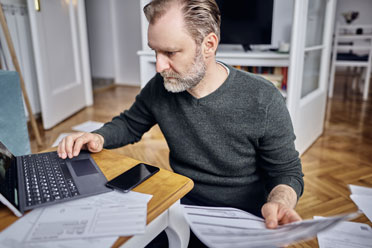 Weißer Mann mittleren Alters arbeitet zuhause am Laptop. In einer Hand hält er ein Formular. Weitere Papiere sind im Zimmer verstreut.