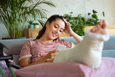 Junge Frau mit gebrochenem Fuß sitzt in einer schonenden Position auf der Couch und schaut lächelnd auf ihr Handy. 