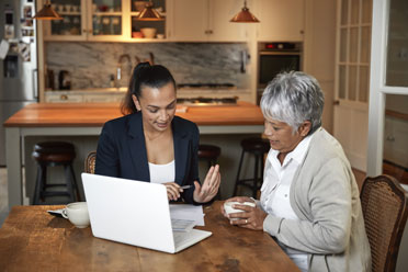10a  Junge Anwältin berät eine Seniorin am Küchentisch im warmen Zuhause der älteren Frau.