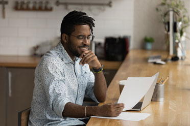 Junger Mann sitzt mit Laptop an der Küchentheke und schaut lächelnd auf ein Blatt Papier. 