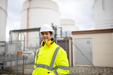 Junger Raffinerie-Arbeiter asiatischer Identität mit gelber Schutzweste, Helm und Kopfhörern, der lächelnd in die Kamera schaut.