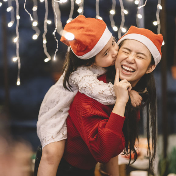 10d-lohnfortzahlung1  Junges Mädchen ostasiatischer Identität auf dem Rücken ihrer großen Schwester. Beide tragen Weihnachtsmützen.