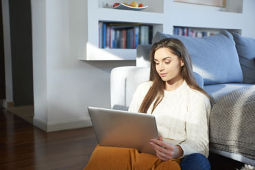 Junge weiße Frau sitzt entspannt auf dem Wohnzimmerboden vor dem Sofa und arbeitet an ihrem Laptop.