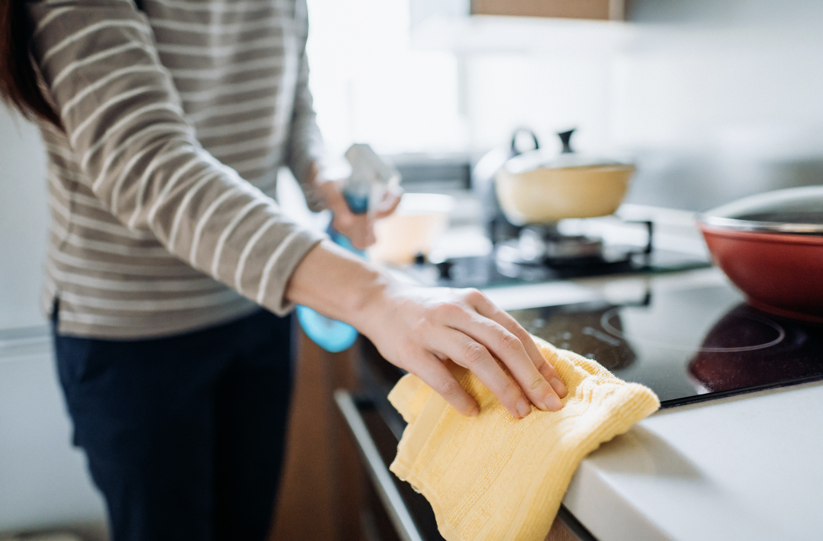 Eine Frau putzt Herdplatte und Küchentheke mit einem gelben Lappen und Reinigungsspray. 