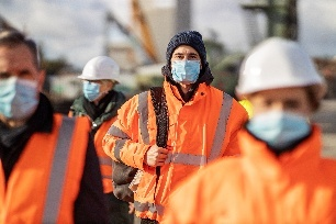 Diverse Gruppe an BauarbeiterInnen mit oranger Schutzweste auf dem Weg nachhause. Fokus liegt auf dem Arbeiter in der Mitte. 