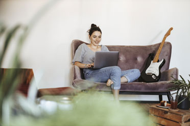 Junge lächelnde weiße Frau sitzt mit Laptop auf dem Schoß zuhause auf ihrer Couch. 
