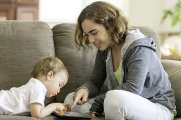 Lächelnde jugendliche Babysitterin spielt mit einem Kleinkind auf dem Sofa im Wohnzimmer.