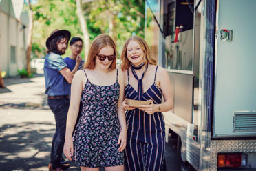 Zwei jugendliche blonde Mädchen entfernen sich lachend von einem Food Truck, an dem sie sich gerade Essen geholt haben. 