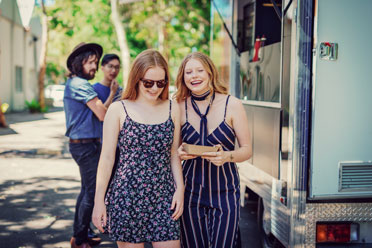 17_Schüler_2  Zwei jugendliche blonde Mädchen entfernen sich lachend von einem Food Truck, an dem sie sich gerade Essen geholt haben.