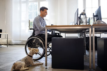 Mann sitzt im Rollstuhl an seinem Schreibtisch in einem Großraumbüro. Neben ihm liegt ein flauschiger Hund. 