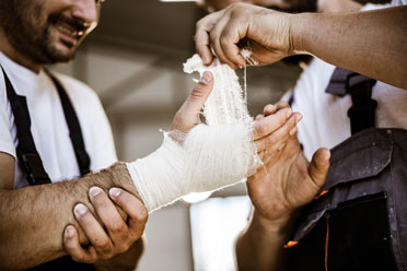 Ein Arbeiter hilft seinem Kollegen dabei dessen verletzte Hand mit einem Verband zu versorgen.