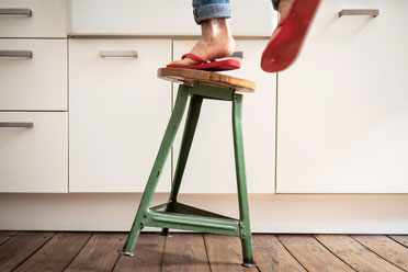 Absicherung bei Unfall1  Eine Person steht auf einem kippelnden Hocker und ist kurz davor herunterzufallen.